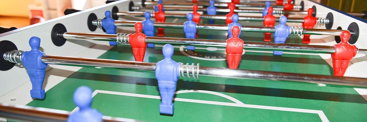 Fussball-Kicker mit grünem Spielfeld, weißer Umrandung und roten und blauen Spielfiguren, die an glänzenden Metallstangen befestigt sind.